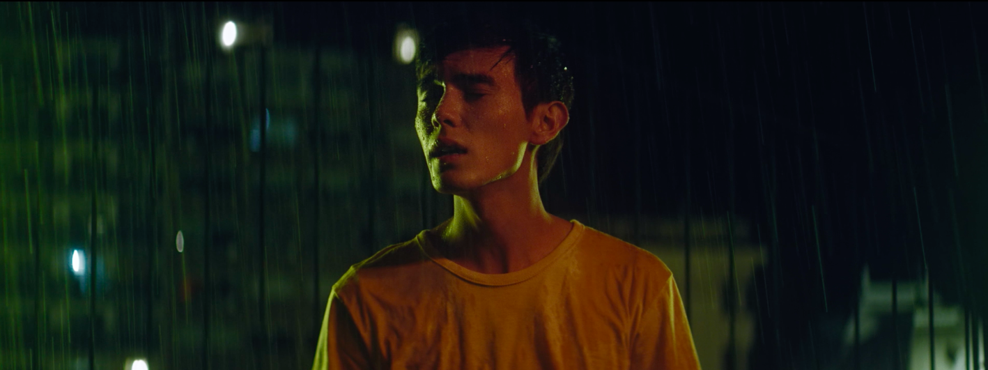 young man with eyes closed and a white tshirt stands in the middle of the street at night, under the rain